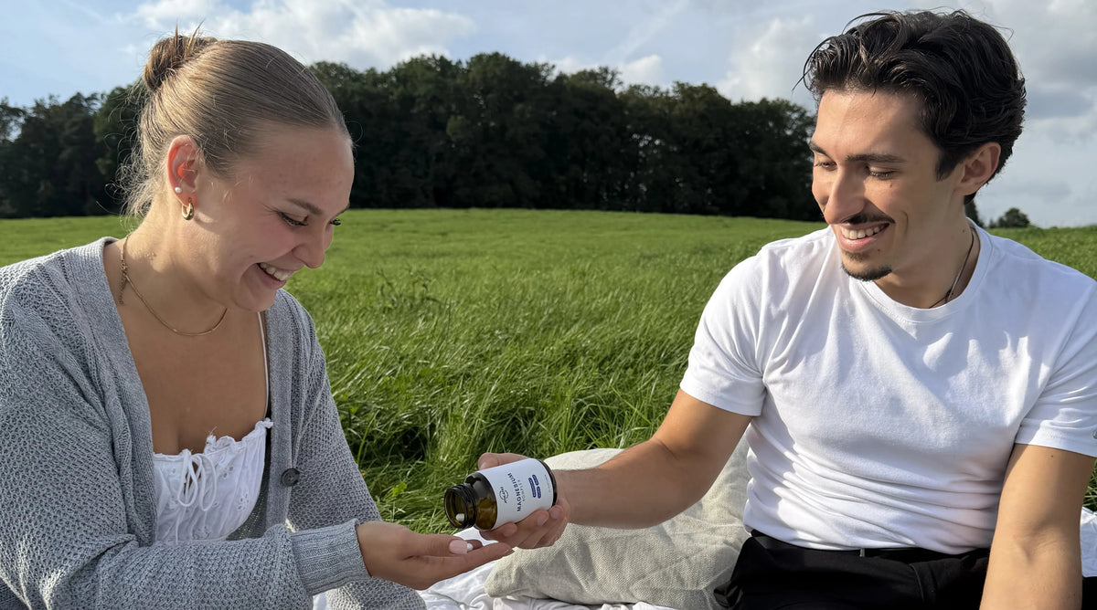 Mann reicht einer Frau ATHLATIQ Magnesium Kapseln beim Picknick auf einer Wiese